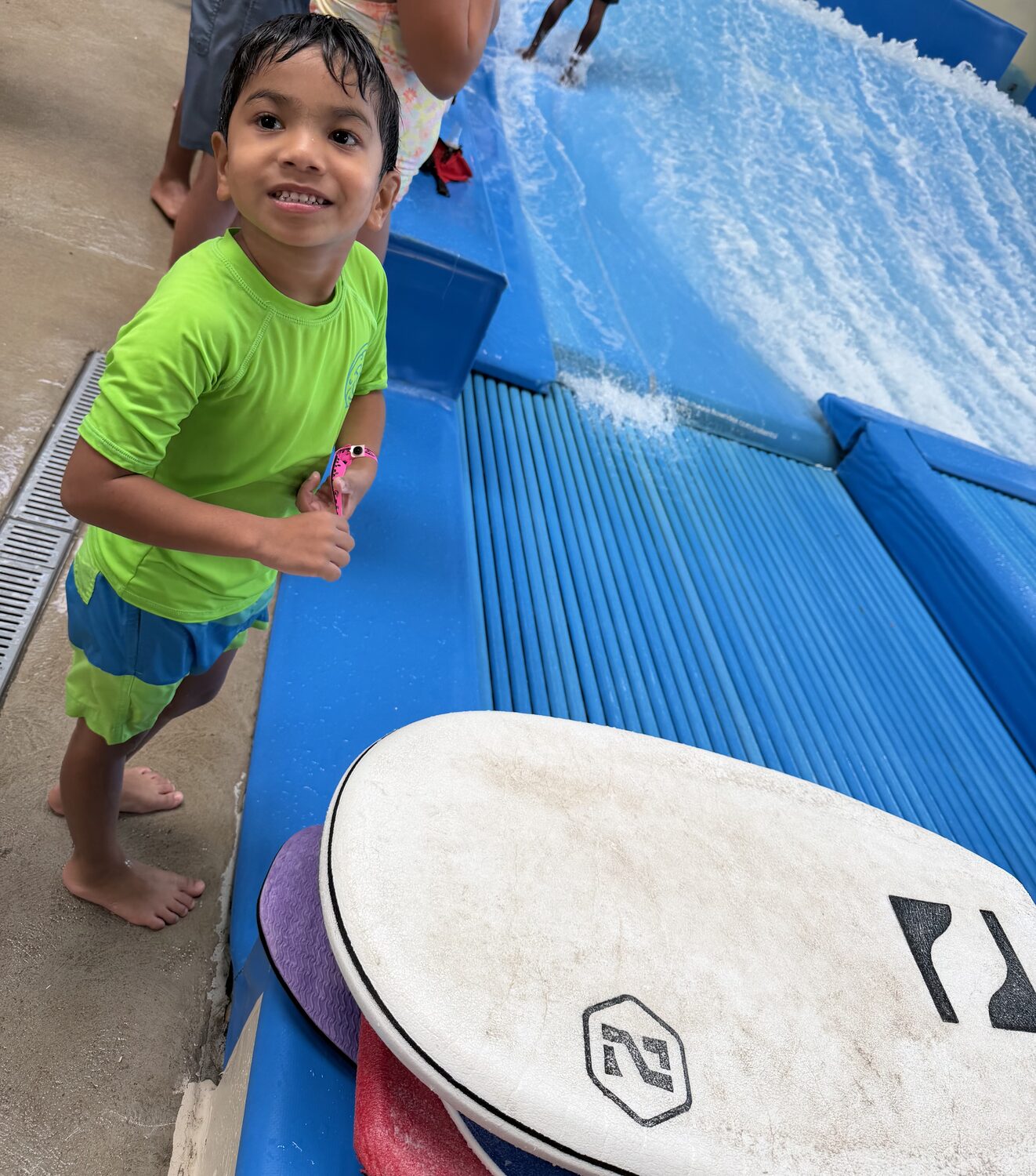 Aadi in a lime green rash guard and blue shorts, hair soaked, grinning at the camera beside a bodyboard at the edge of the FlowRider, with the wave rushing behind him.