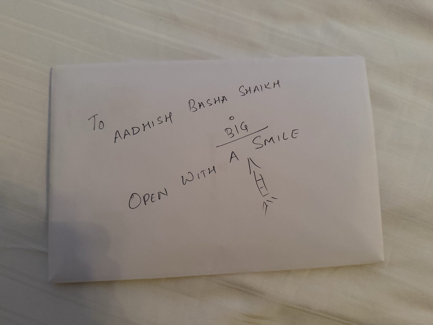 A white envelope handwritten with 'To Aadhish Basha Shaikh, open with a BIG smile' and a small arrow doodle.