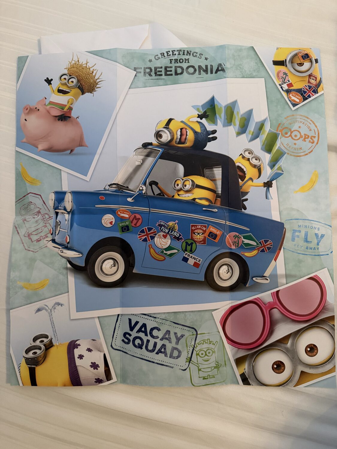 Minions-themed wrapping paper with a blue van covered in travel stickers, reading 'Greetings from Freedonia' and 'Vacay Squad'.